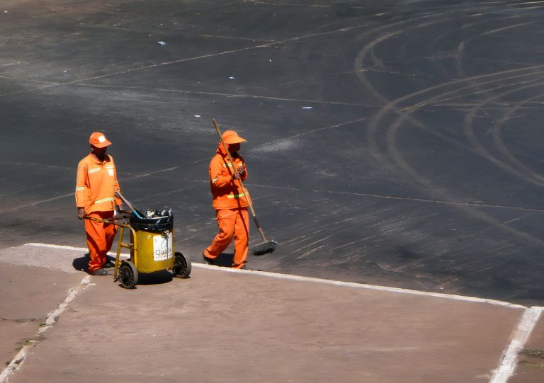 Dois trabalhadores de limpeza urbana usam uniformes laranja enquanto trabalham em uma área pública de Brasília.